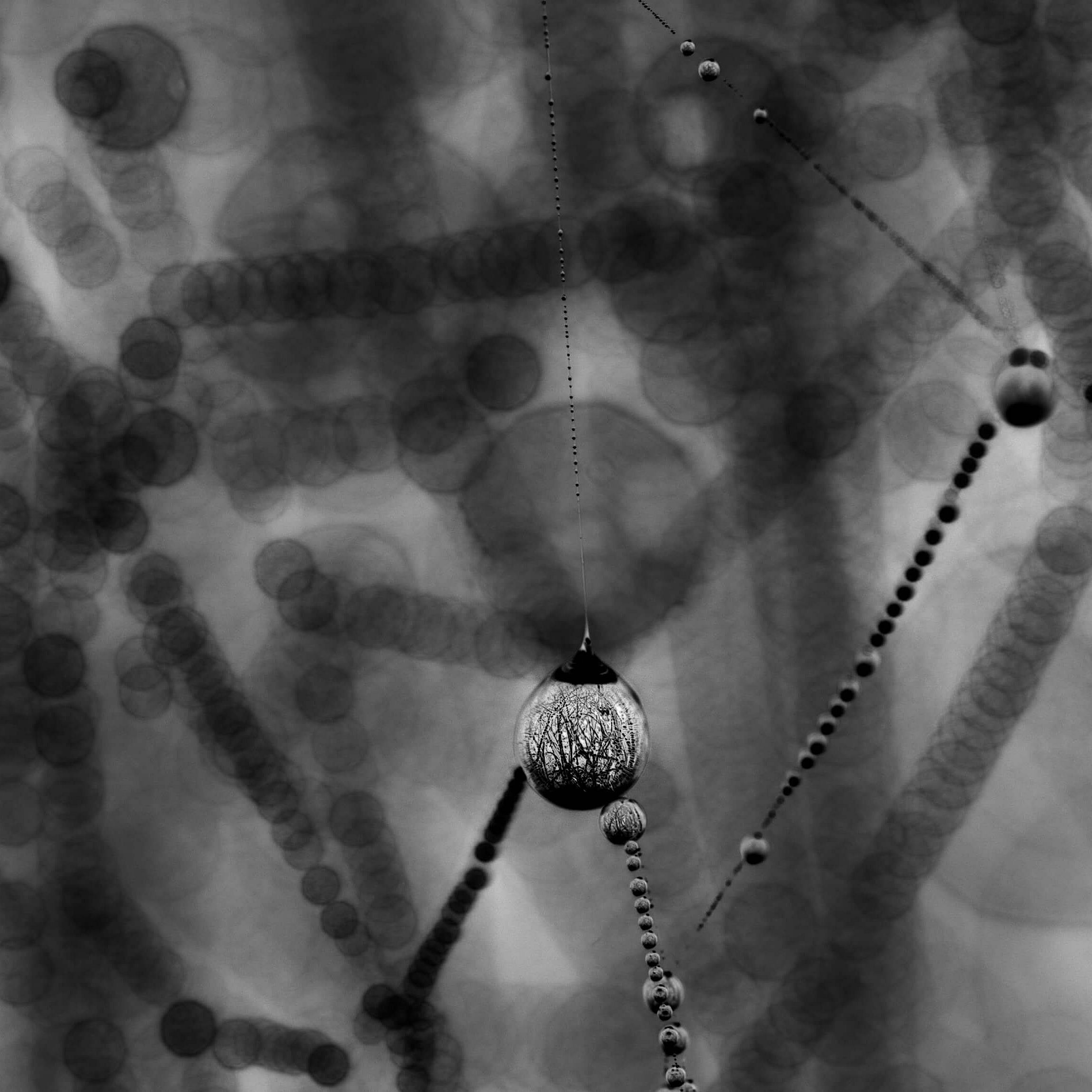 Ein unscharfes, monochromes Bild mit vielen kreisförmigen Formen, die in verschiedenen Größen und Hell-Dunkel-Kontrasten angeordnet sind. Im Vordergrund hängt eine auffällige, runde Perle oder Kugel, die durch ihre Textur hervorsticht. Die Umgebung wirkt abstrahiert und verschwommen.