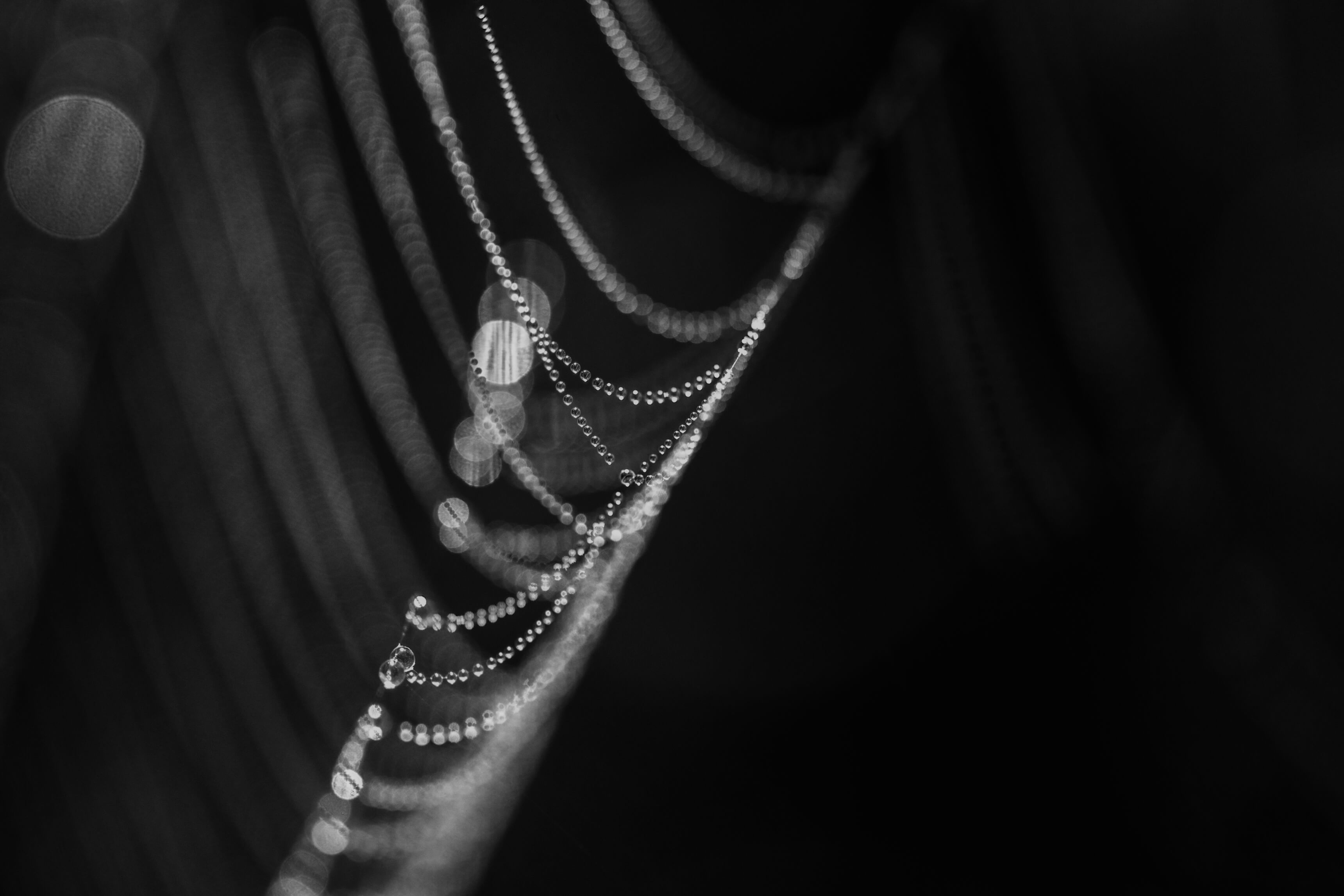Ein feines Spinnennetz, mit Wassertropfen bedeckt, zeigt glitzernde Perlen in Schwarz-Weiß. Die Tropfen reflektieren das Licht und erzeugen eine zarte, schimmernde Atmosphäre. Das Netz ist kunstvoll aufgefädelt und zieht sich diagonal über den Hintergrund.